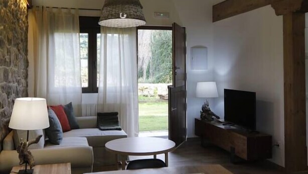 Una sala de estar acogedora con pared de piedra, techo de madera, un sofá, un mueble para televisión, una mesa y una lámpara de techo grande de fibras naturales.