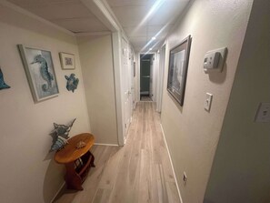 A hallway with wooden flooring, a small round table with a cushion, and wall-mounted artwork including seahorse and floral designs.