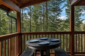 Une table ronde avec quatre assiettes bleues et deux chaises, placée sur une terrasse en bois offrant une vue sur une forêt.