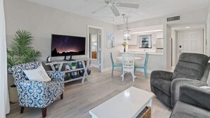 A modern living room with a flat-screen TV, patterned armchair, grey sofa, and a dining area with a white table and chairs. - F-338 -Beautiful Resort and Sunset Views! (Naples)