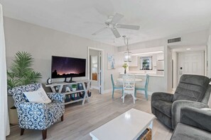 A modern living room with a flat-screen TV, patterned armchair, grey sofa, and a dining area with a white table and chairs. - F-338 -Beautiful Resort and Sunset Views! (Naples)
