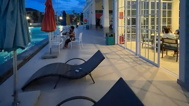 An outdoor pool area with lounge chairs and umbrellas, a covered seating area with tables, and a building with large glass doors.