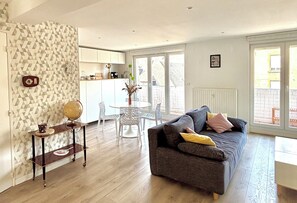 Un salon avec un canapé gris, une petite table ronde et une chaise blanche. On y trouve également un coin cuisine avec des meubles blancs et une fenêtre.