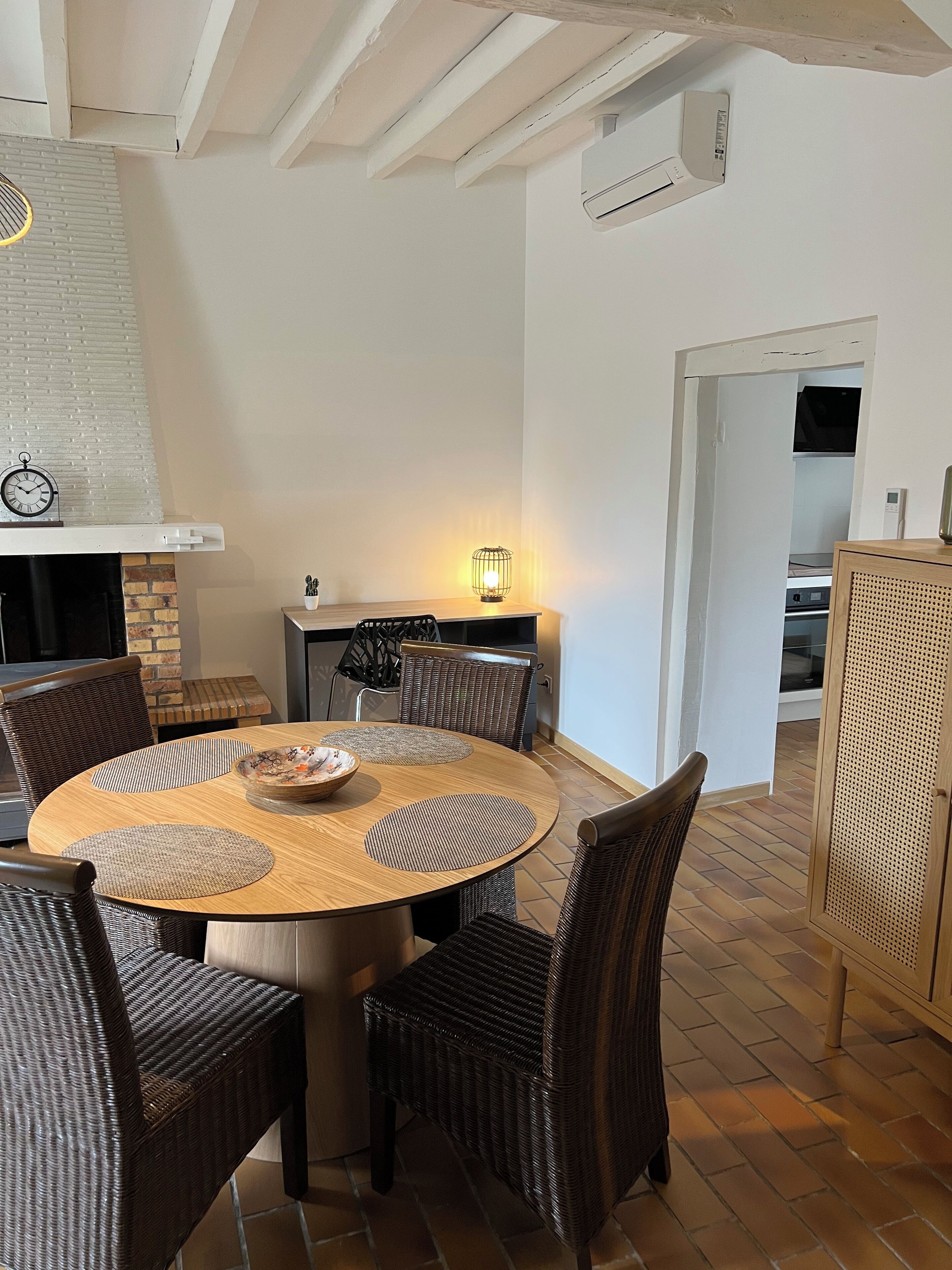 A dining area with a round wooden table, wicker chairs, a wall-mounted clock, and a lit wall lamp.