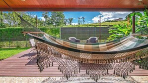 A hammock with chairs underneath, set on a wooden deck with a view of a green landscape and a house in the background. - Casa Heliconias La Fortuna (La Fortuna)