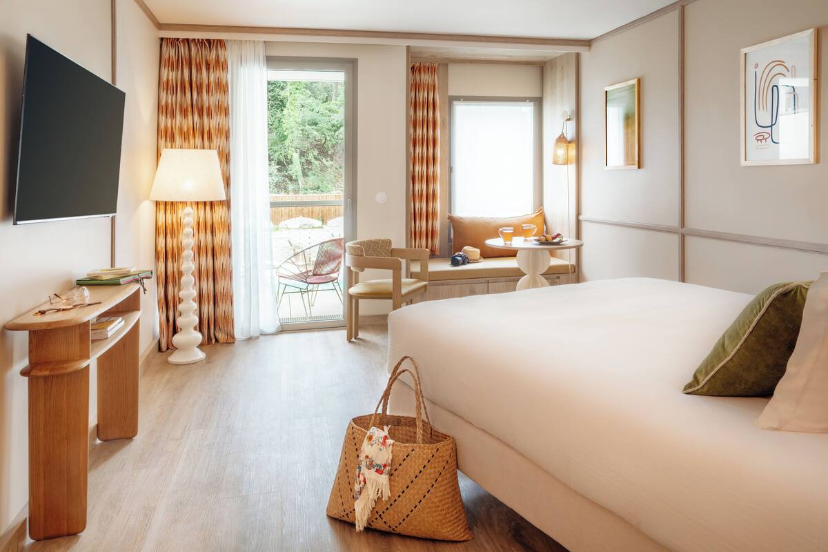 Ein Hotelzimmer mit einem Bett, einem Schreibtisch, einem Sessel, einem Tisch mit Getränken, einem Fernseher und einem Fenster mit Vorhängen.