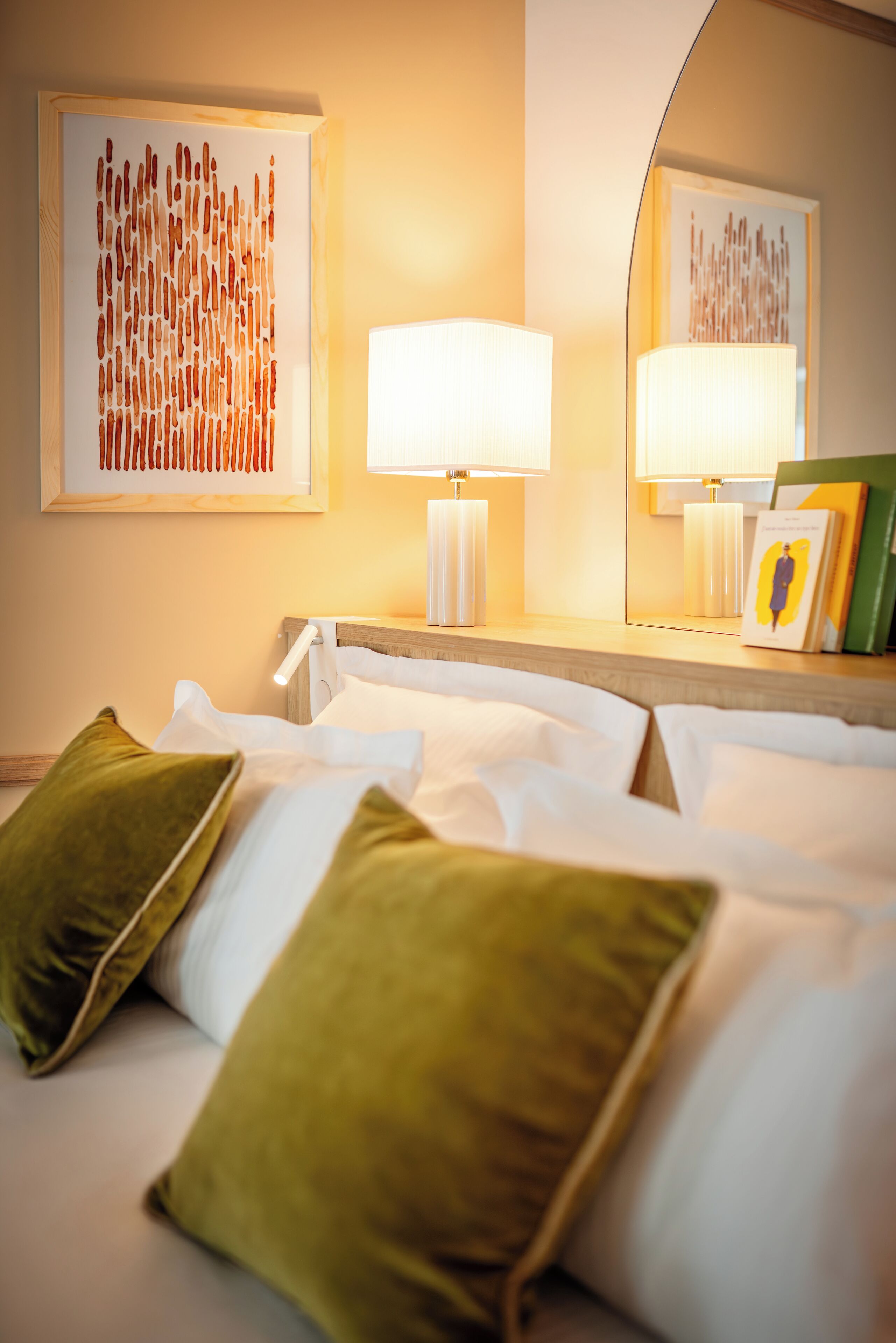 A hotel room with a neatly made bed, two green pillows, a bedside lamp, and two framed artworks on the wall.