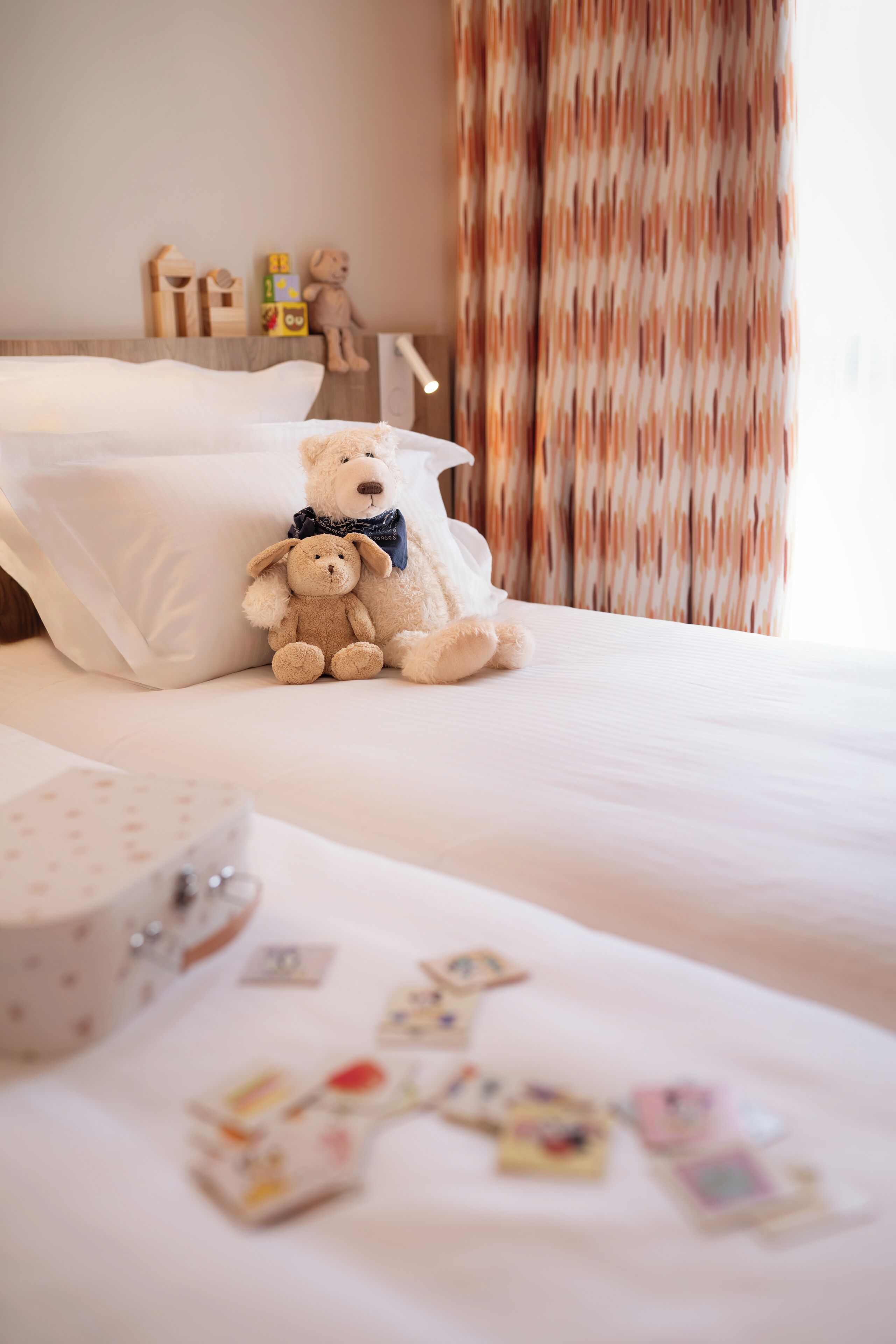 A neatly made bed with two teddy bears, a wooden toy set, and a curtain.