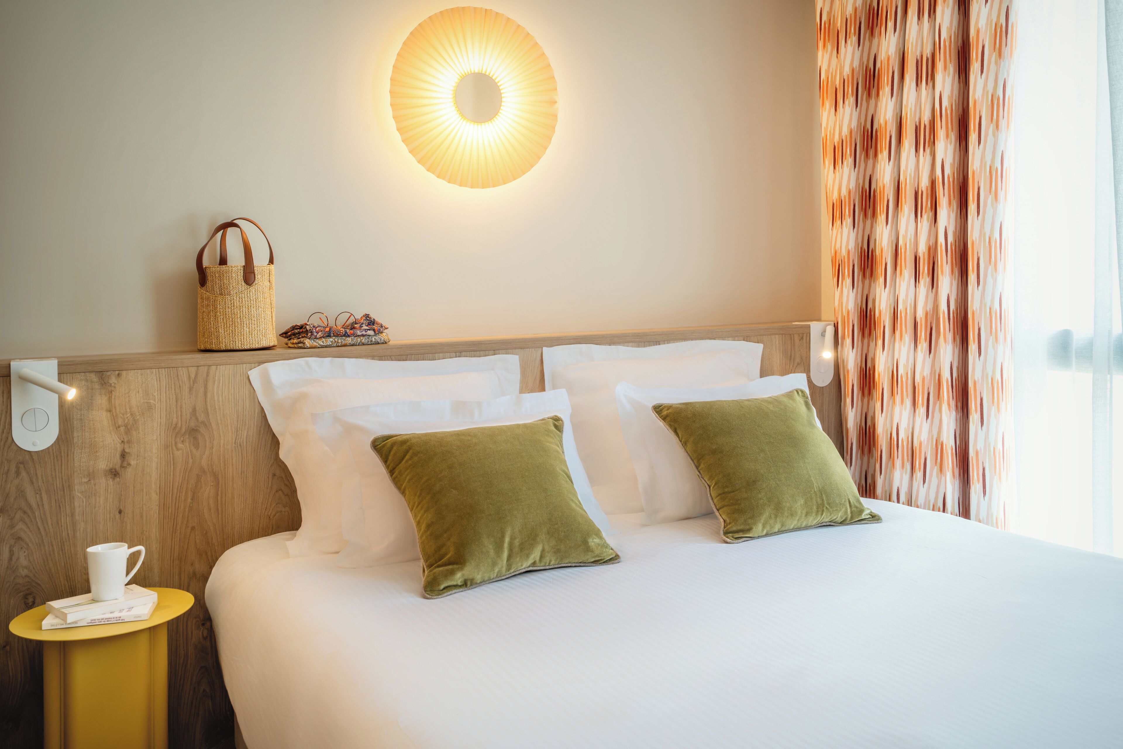 Une chambre d’hôtel avec un lit, des oreillers, une table de chevet avec une tasse et des livres, une lampe fixée au mur et un rideau.