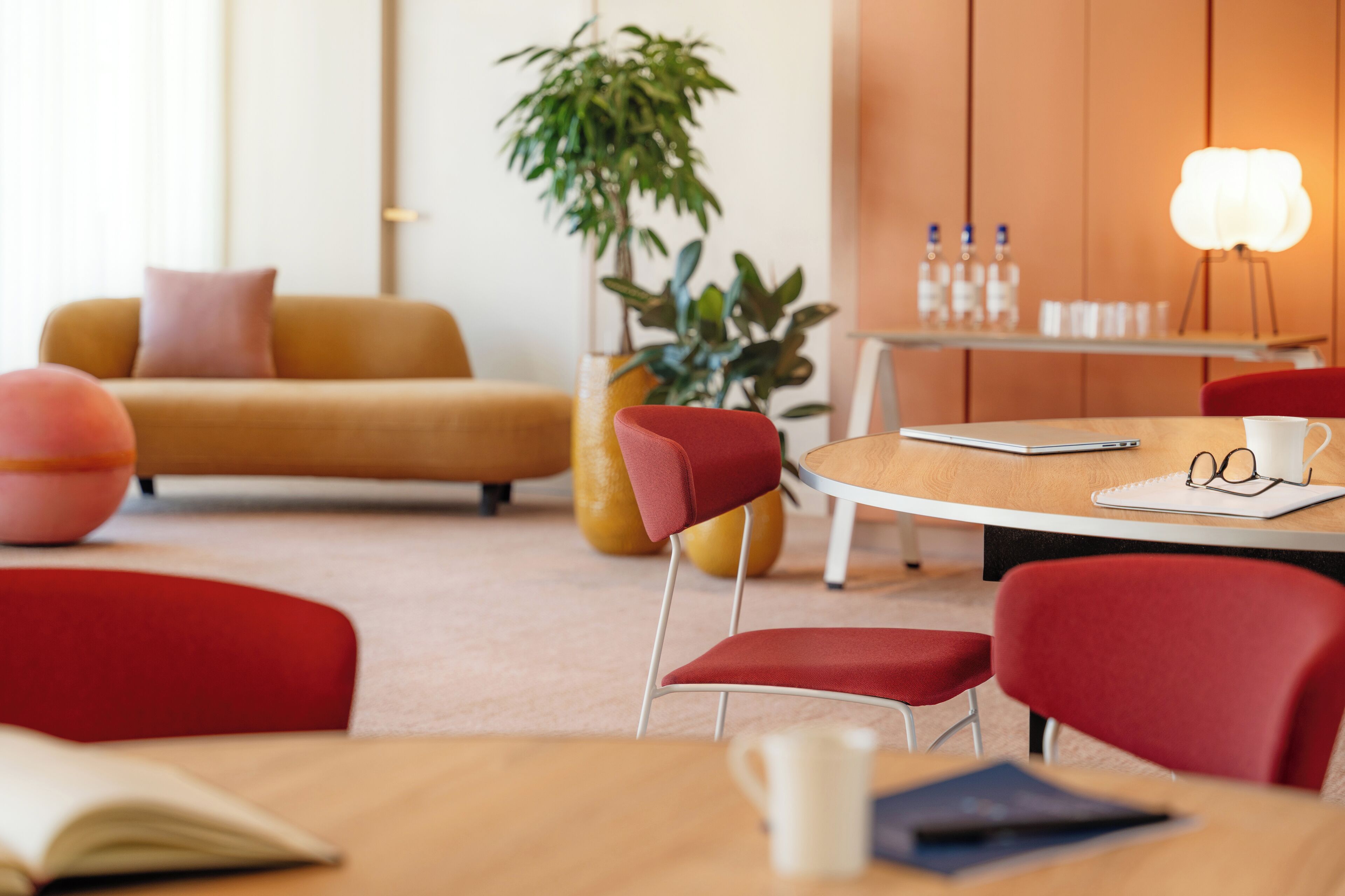 Une chambre d’hôtel moderne avec une table ronde, des chaises rouges, un canapé et une plante en pot.