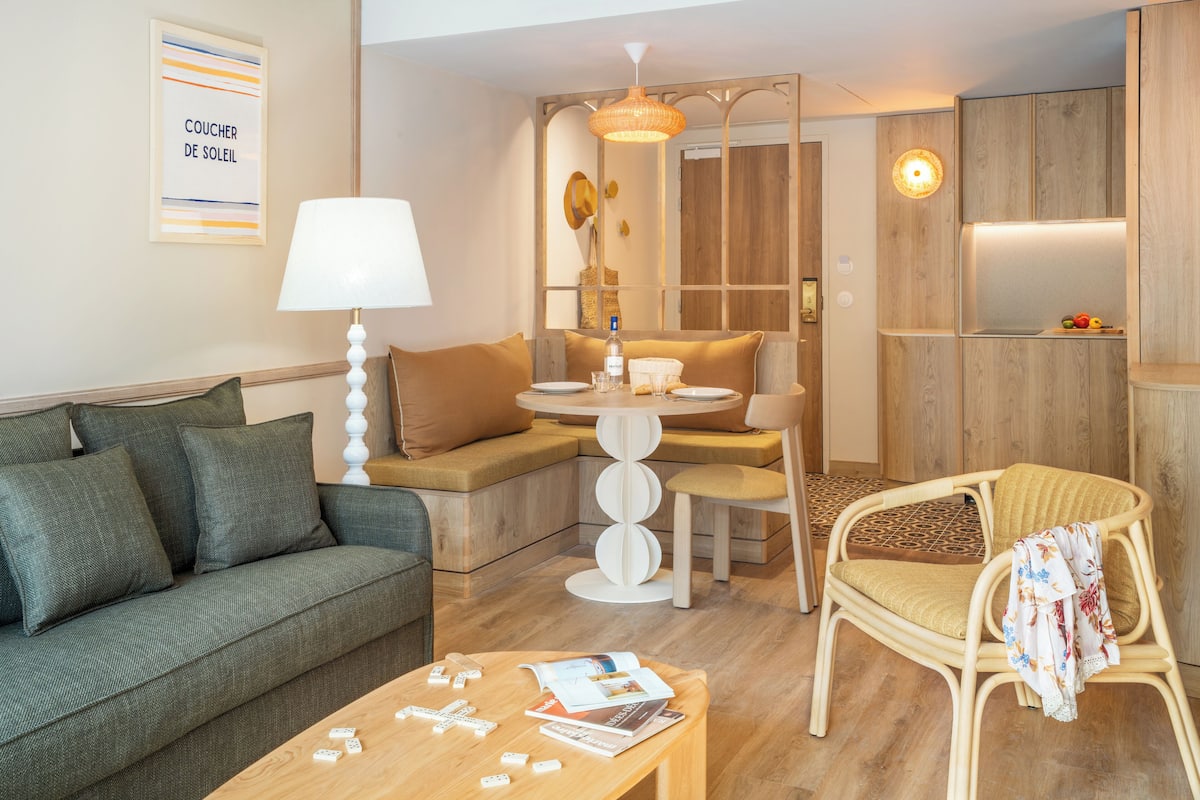 Ein modernes Wohnzimmer mit einer Couch, einem Esstisch und einem Sessel. Eine Lampe, ein Beistelltisch mit Zeitschriften und einer Fernbedienung sowie eine Wand mit einem gerahmten Bild sind zu sehen.