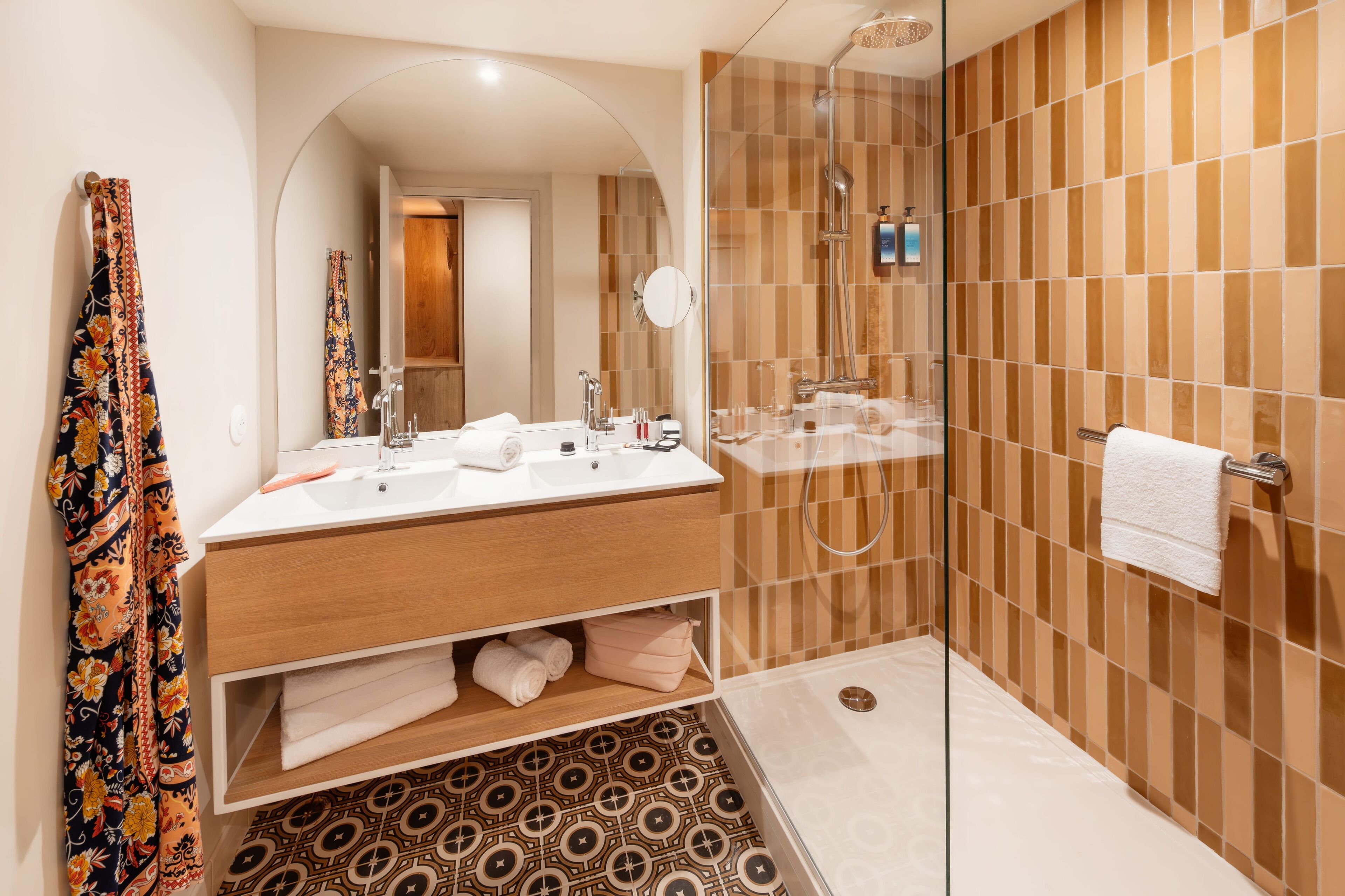 Un baño moderno con ducha envuelta en vidrio, tocador de madera y baldosas con diseño en el suelo.