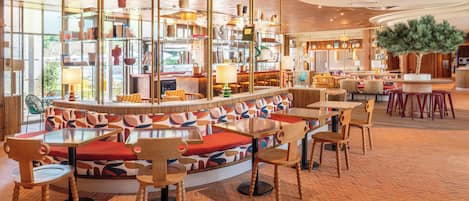 Un restaurante moderno con una zona central de estar que cuenta con un banco circular con cojines de colores, rodeado de mesas y sillas de madera.