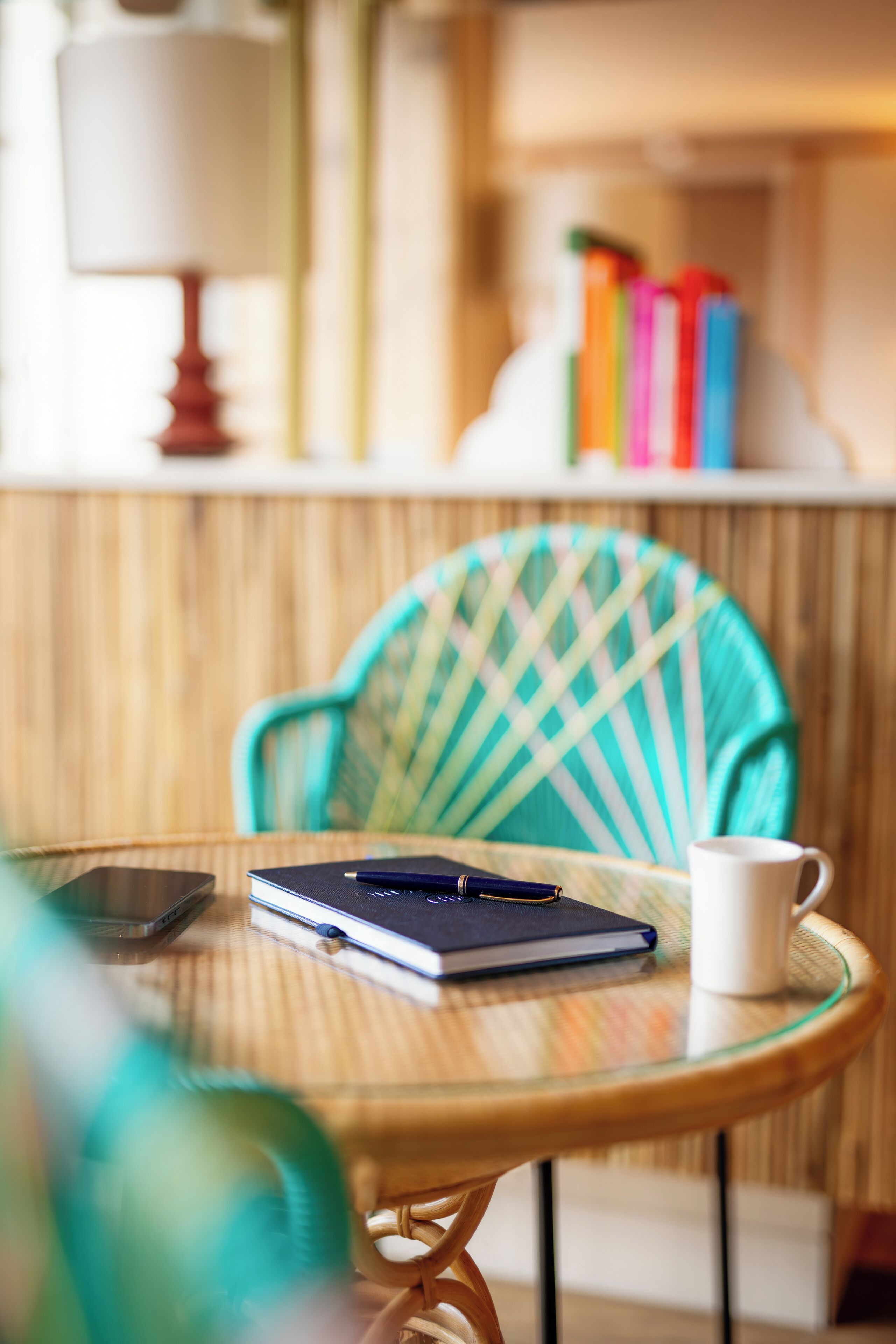 A wicker chair with a checkered cushion, a glass table with a notebook, pen, and a white mug.