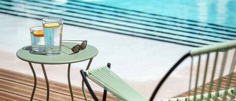 Zona de piscina con una silla, una mesita con vasos y un limón, y un bolso a rayas sobre una terraza de madera.