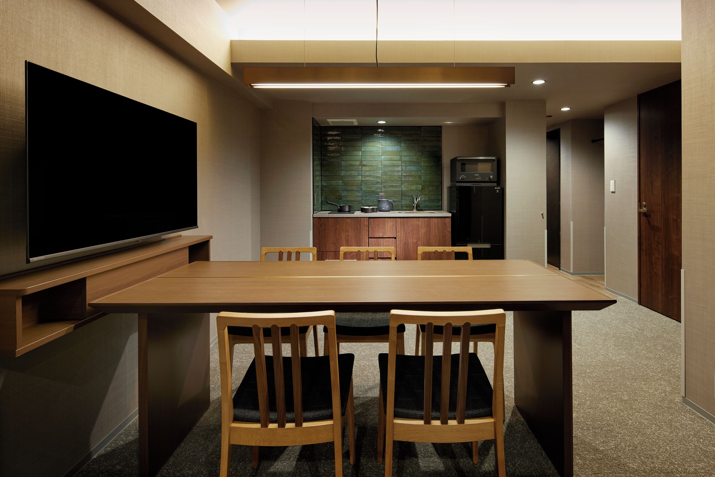 Habitación de hotel moderna con un televisor de pantalla plana grande montado en la pared, una mesa de comedor de madera con cuatro sillas, una zona de kitchenette con microondas y un refrigerador pequeño, y un piso cubierto de alfombra oscura.