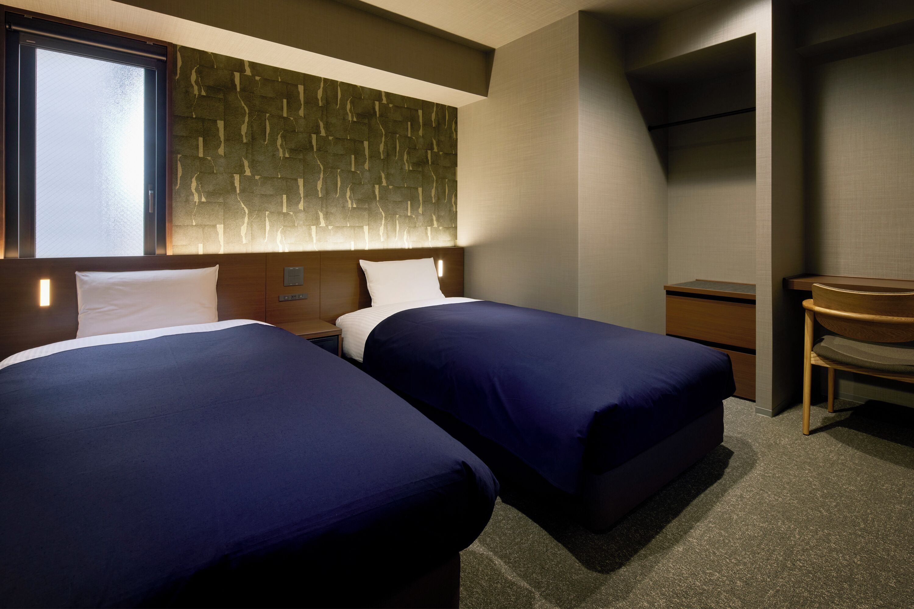 A hotel room with two beds, a wooden headboard, a small desk, and a chair.