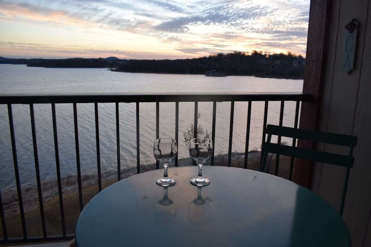 A balcony with a table and two glasses, overlooking a body of water with a sunset view.
