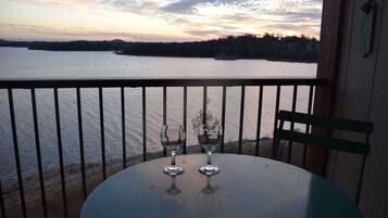 A balcony with a table and two glasses, overlooking a body of water with a sunset view.