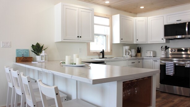 A modern kitchen with white cabinetry, a central island with bar stools, stainless steel appliances, and a window providing natural light. - Newly Built Ocean View Cottage, Minutes To Thunder Cove Beach (Kensington)