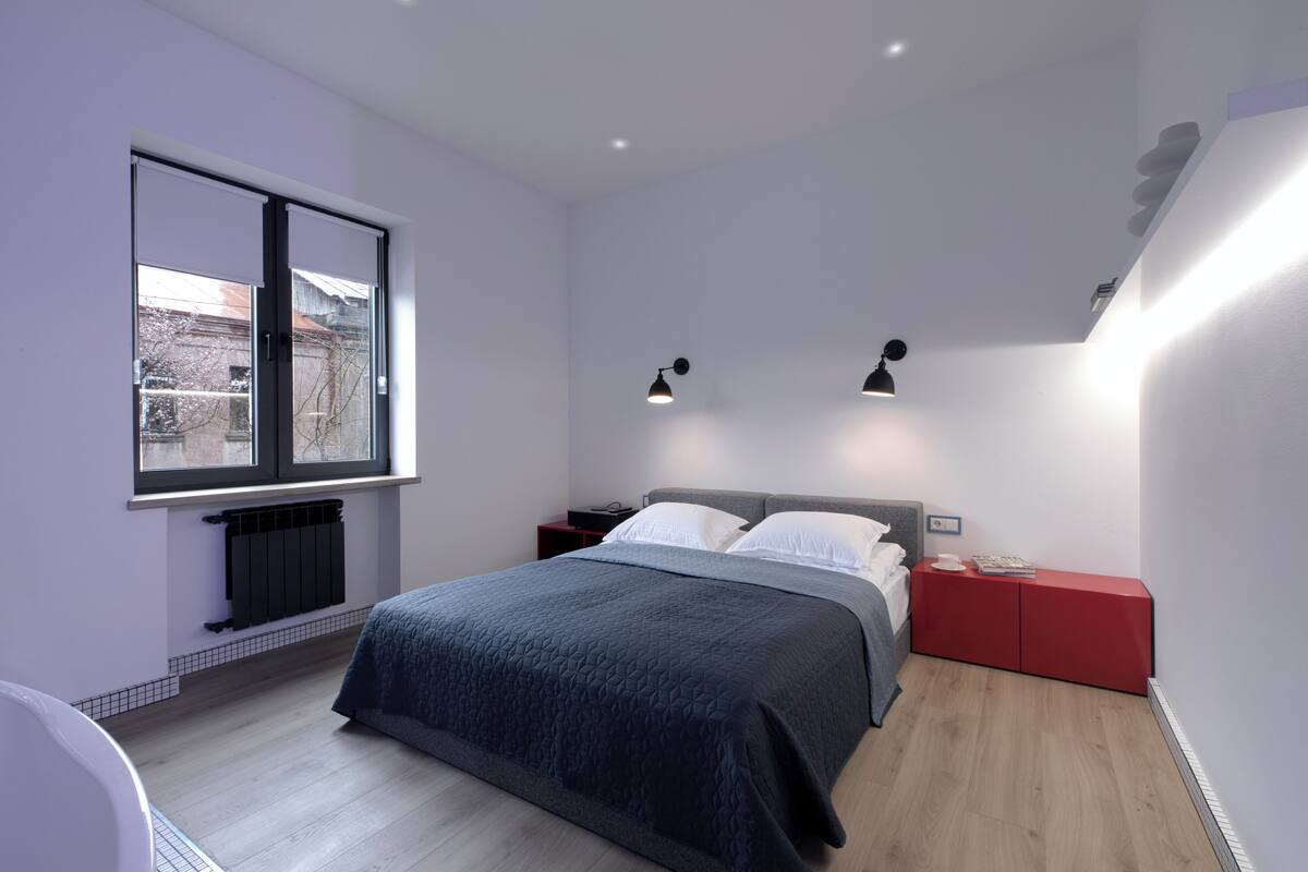 A modern bedroom with a large window, a bed with a dark quilt, two bedside tables with lamps, and a red nightstand.