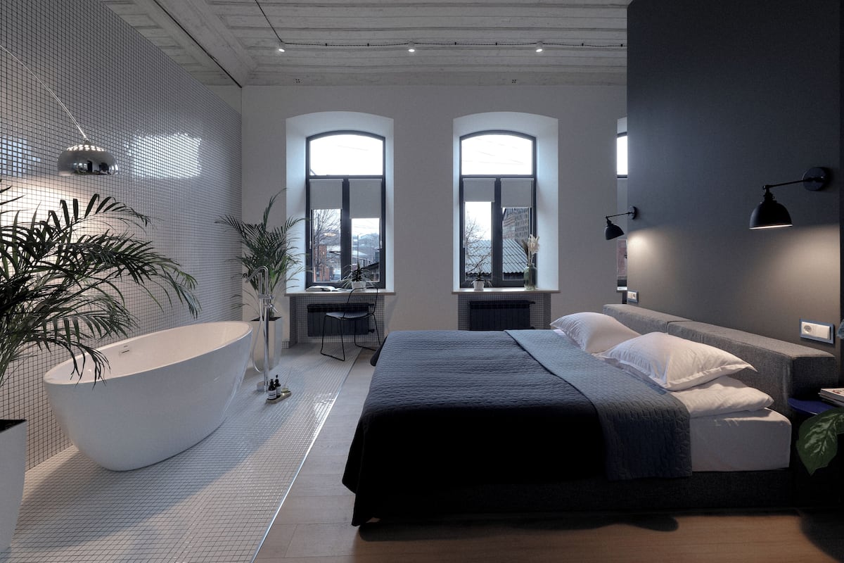 A modern bedroom with a large bathtub, two windows, a desk, and a bed with a dark-colored bedspread.