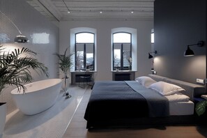 Une chambre à coucher moderne avec une grande baignoire, deux fenêtres, un bureau et un lit recouvert d’une couvre-lit de couleur sombre.
