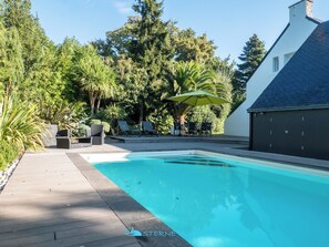 A swimming pool with lounge chairs and an umbrella, surrounded by lush greenery and a white building with a dark roof. - Superb house 100m from the sea with heated swimming pool (Concarneau)