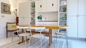 A modern kitchen with white cabinets, a wooden dining table, and four white chairs. There is a sofa with a green cushion in the background.