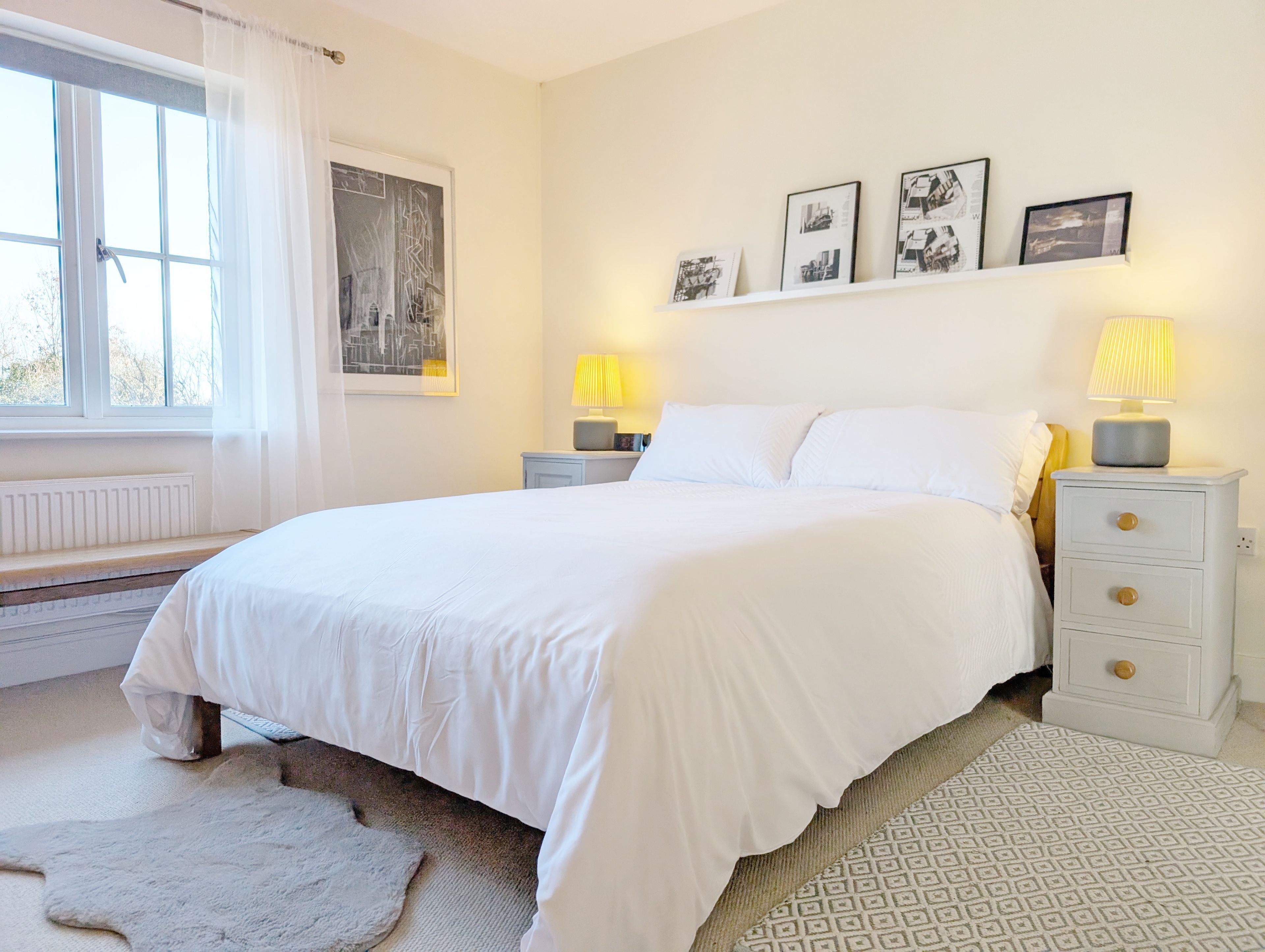 A bedroom with a large bed, two bedside tables with lamps, a radiator, a window with curtains, and a wall with framed pictures.