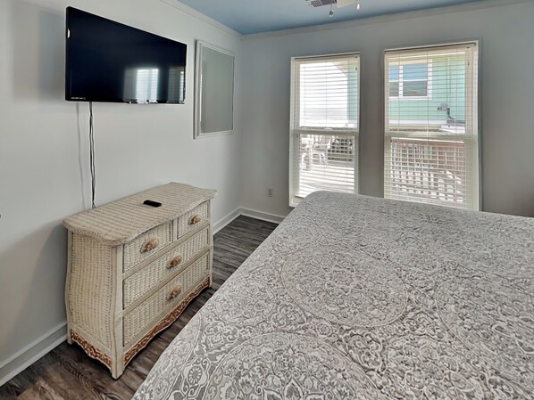 A bedroom with a large bed, a television mounted on the wall, a wicker chest of drawers, and a balcony with a view of a house. - Once Upon A Time | A Spacious Beachfront (Galveston)