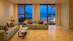 A spacious living room with a large window offering a view of the beach and ocean at dusk. There are two sofas with colorful cushions and a small table in the center.