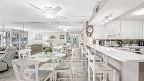 Una cocina moderna con armarios blancos, una isla central y un comedor con sillas blancas y una mesa de vidrio.