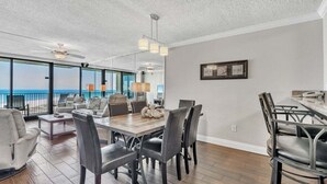 A spacious dining area with a large wooden table and modern chairs, overlooking a balcony with a view of the beach.