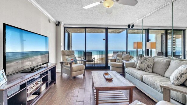 A living room with a large TV, comfortable seating, and a balcony with a view of the beach.