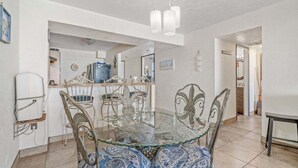 Un comedor con una mesa de vidrio rodeada de sillas, una cocina al fondo y una puerta que da a otra habitación.