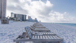 Una playa con sillas de madera para tomar el sol alineadas, arena y edificios altos al fondo.