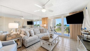 A spacious living room with a large sofa, a coffee table, and a television. There is a balcony with a view of the ocean and a sailboat model.