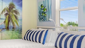 Una camera da letto con un letto, cuscini a righe, un quadro con palme e una finestra con persiane.