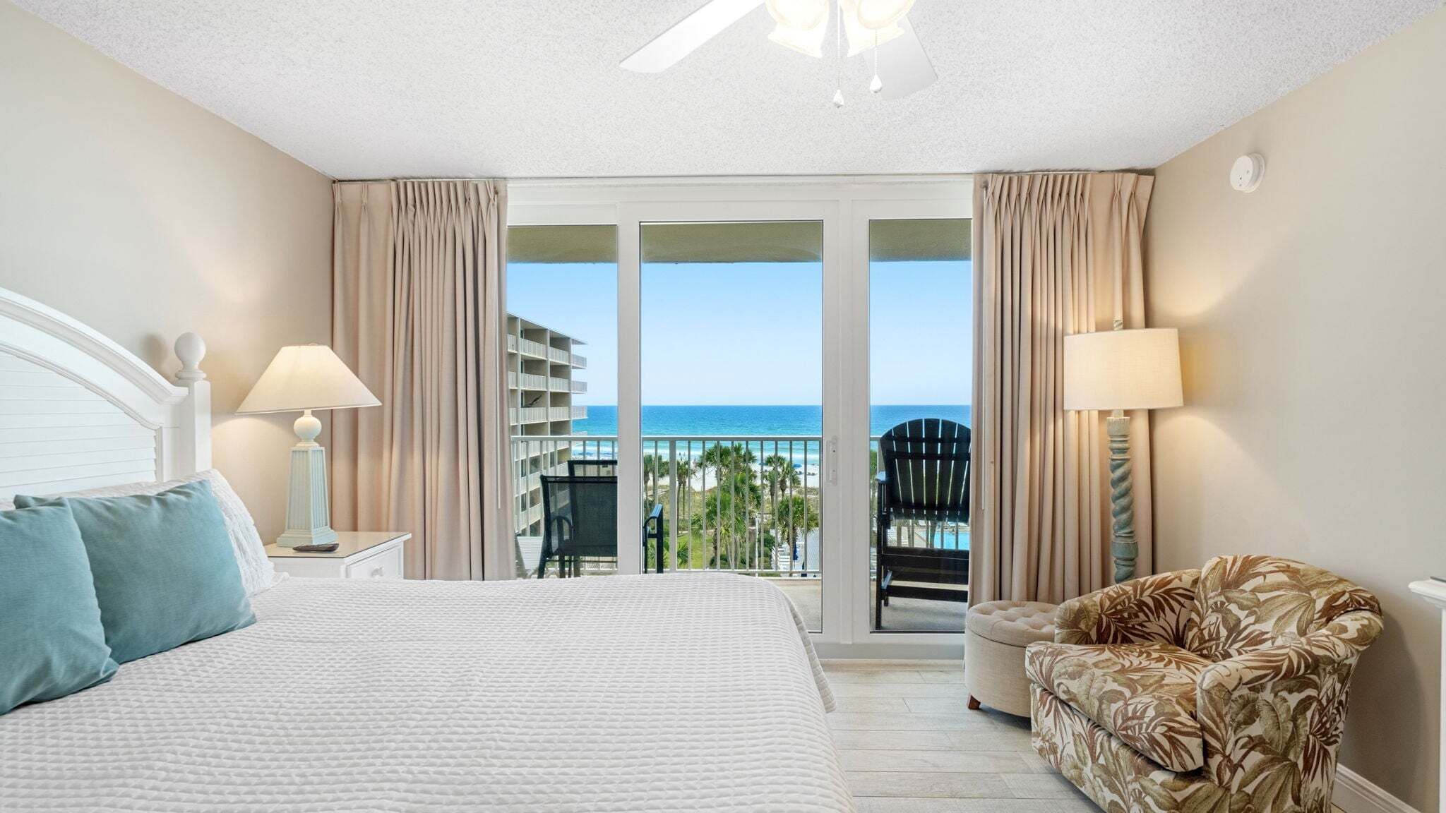 A hotel room with a bed, two bedside lamps, a chair, and a balcony with a view of the ocean and a building.