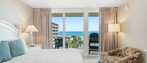 A hotel room with a bed, two bedside lamps, a chair, and a balcony with a view of the ocean and a building.