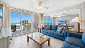 A modern living room with a blue sofa, a glass coffee table, and a TV. There is a balcony with a view of the beach and buildings.