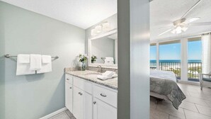 A bathroom with a large mirror, a sink, and a towel rack. There is a balcony with a view of the ocean.