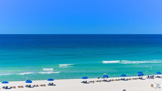 A beach with white sand, clear turquoise water, and a boat in the distance. There are blue umbrellas and lounge chairs along the shore.
