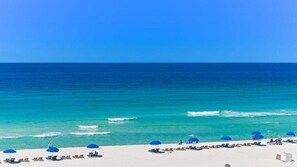 A beach with white sand, clear turquoise water, and a boat in the distance. There are blue umbrellas and lounge chairs along the shore. - Dunes C504-King Bed-Beachfront-Pet Friendly (Panama City Beach)