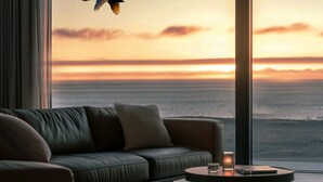 Un salon moderne avec un canapé en cuir, une table basse ronde et une vue sur l’océan au coucher du soleil.