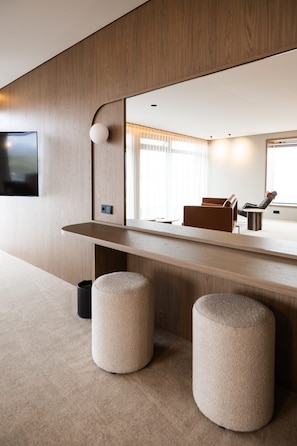 A modern hotel room with a wooden accent wall, a flat-screen TV mounted above a wooden desk, and a large mirror reflecting the room.