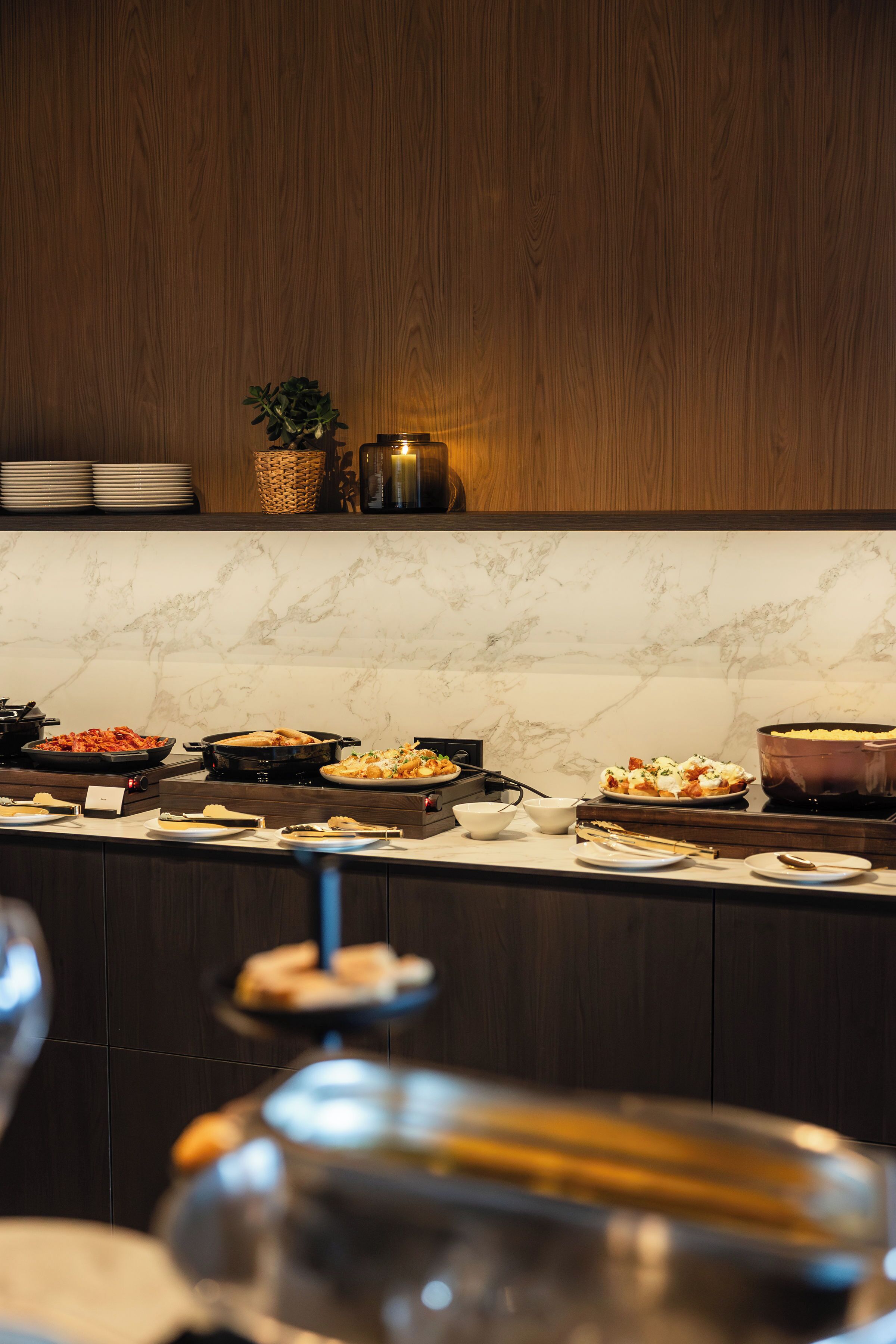Disposição de buffet com diversos pratos, uma vela e uma planta sobre uma estante de madeira.