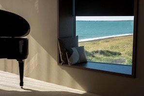 A piano in a room with a window overlooking a grassy area and the sea. - Black Sand Hotel (Sveitarfélagid Ölfus)