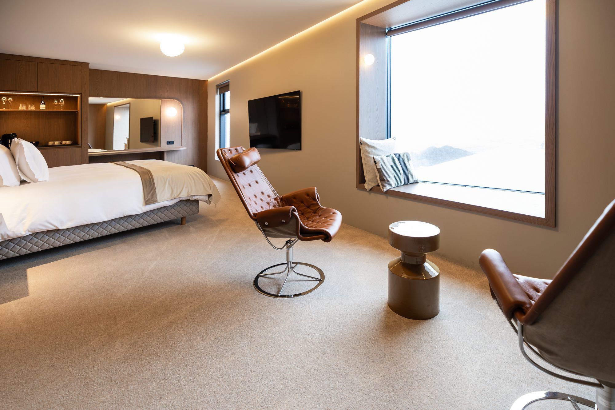 A modern hotel room with a bed, a seating area with a chair, a television, and a window with a view.